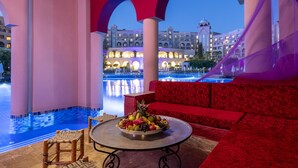 A luxurious outdoor seating area with a pool view, a fruit bowl on a table, and a red patterned sofa.