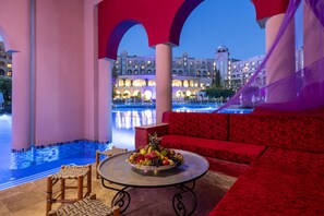 A luxurious outdoor seating area with a pool view, a fruit bowl on a table, and a red patterned sofa.
