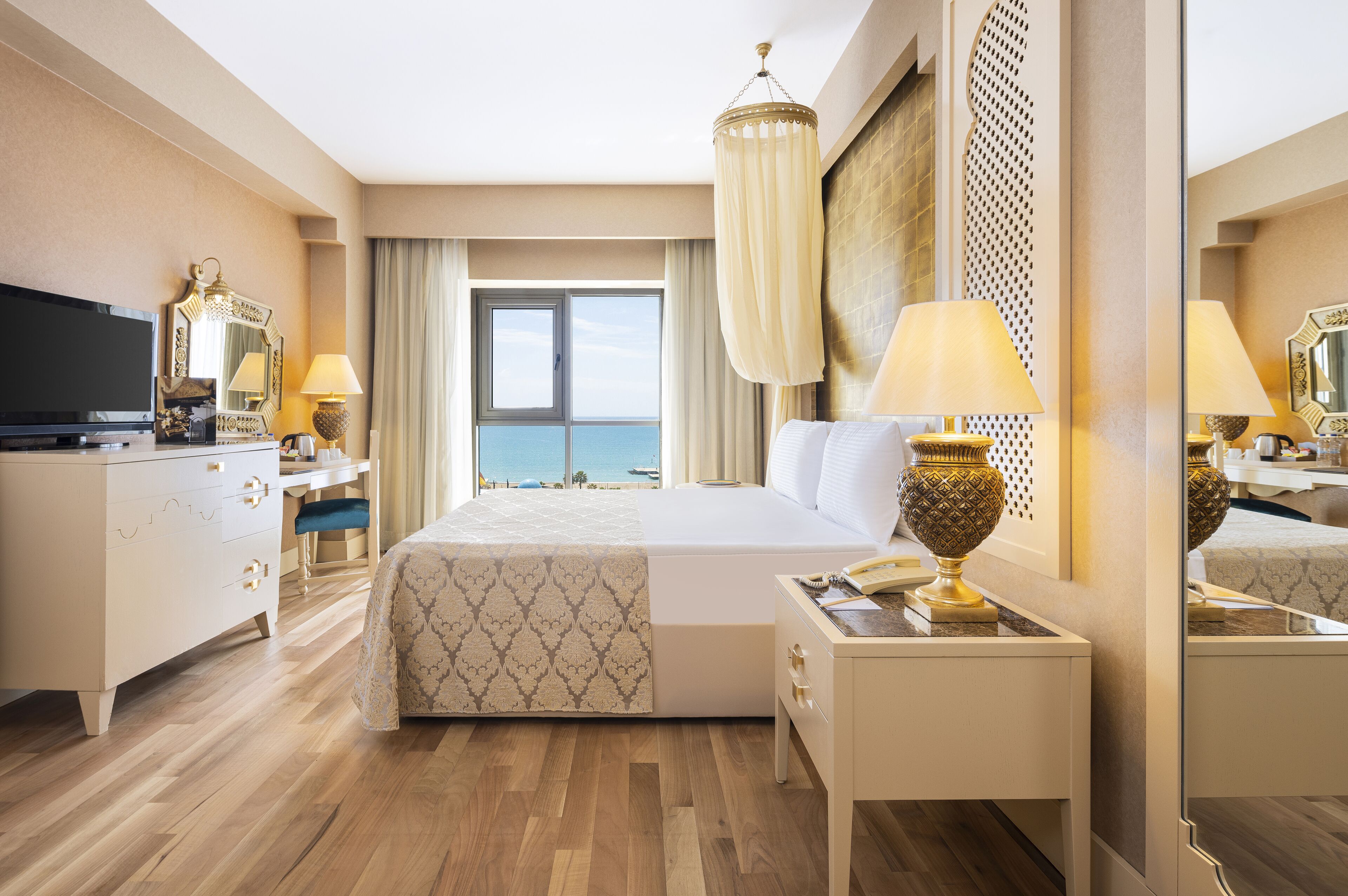 A hotel room with a large bed, a desk with a TV, a lamp, and a view of the sea through the window.