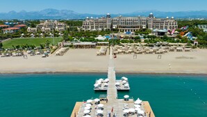 Et hotel ved vandet med en mole, der strækker sig ud i havet, med udendørs siddepladser og udsigt til en strand og bjerge i baggrunden.