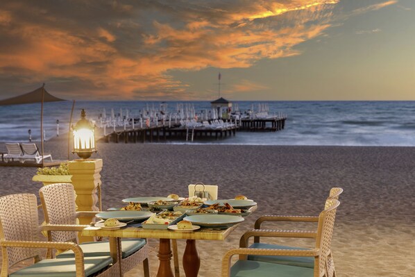 Montaje de comedor junto a la playa con sillas de mimbre y una mesa dispuesta para una comida, con vistas al océano y un muelle.