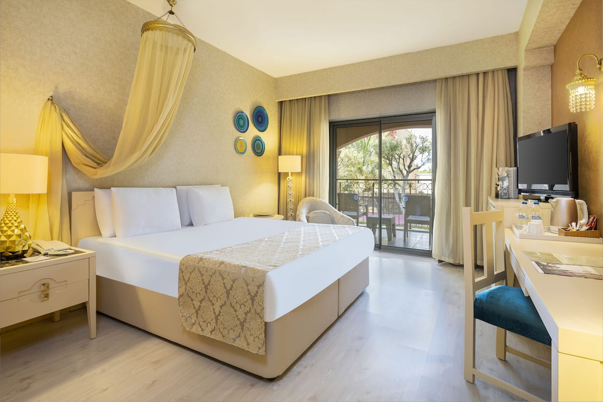 Ein Hotelzimmer mit einem großen Bett, einem Schreibtisch mit Stuhl, einem Fernseher und einem Balkon mit Vorhängen.