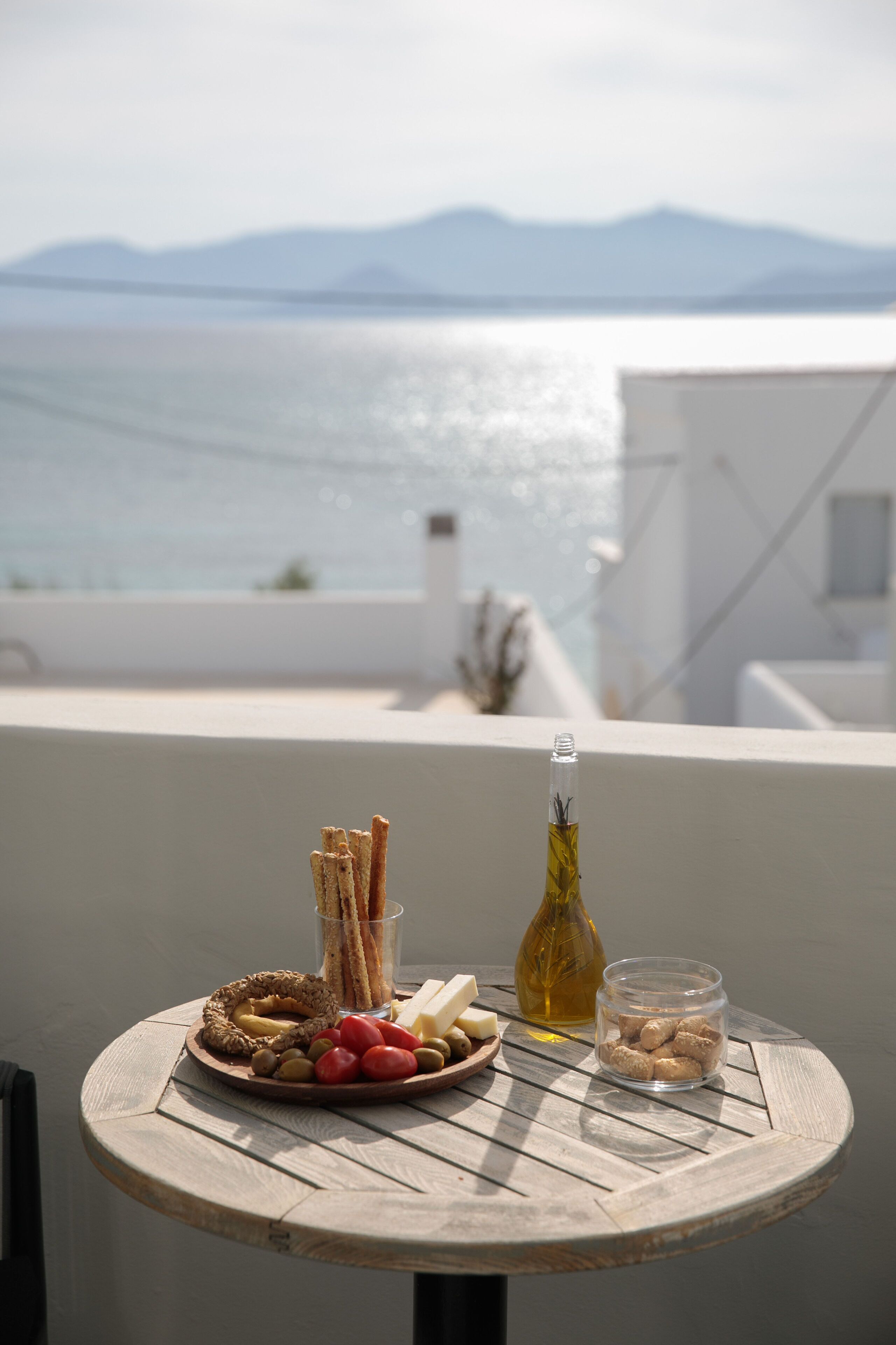 A round wooden table with a plate of snacks, a glass of water, and a bottle of olive oil on a balcony overlooking a body of water and mountains.