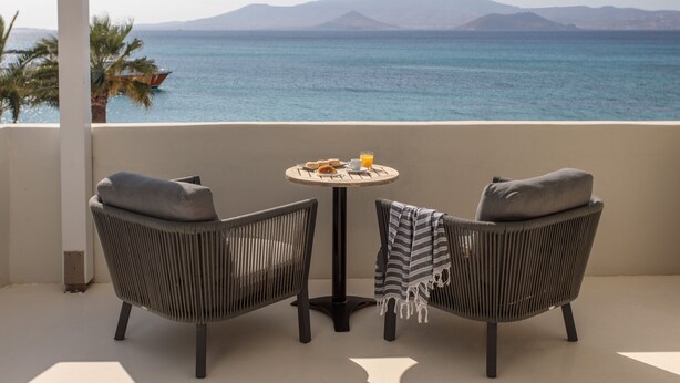 A balcony with two wicker chairs, a small round table with breakfast items, and a view of the sea and mountains. - Iria Beach art hotel (Naxos)