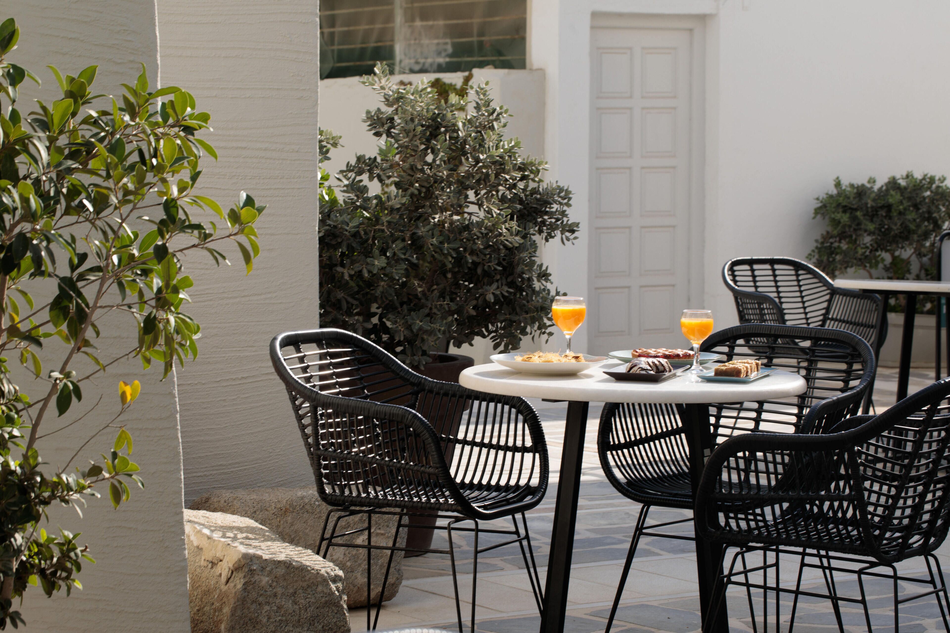 Een terras met een ronde tafel en vier zwarte stoelen, omgeven door plantenbakken en een witte deur.