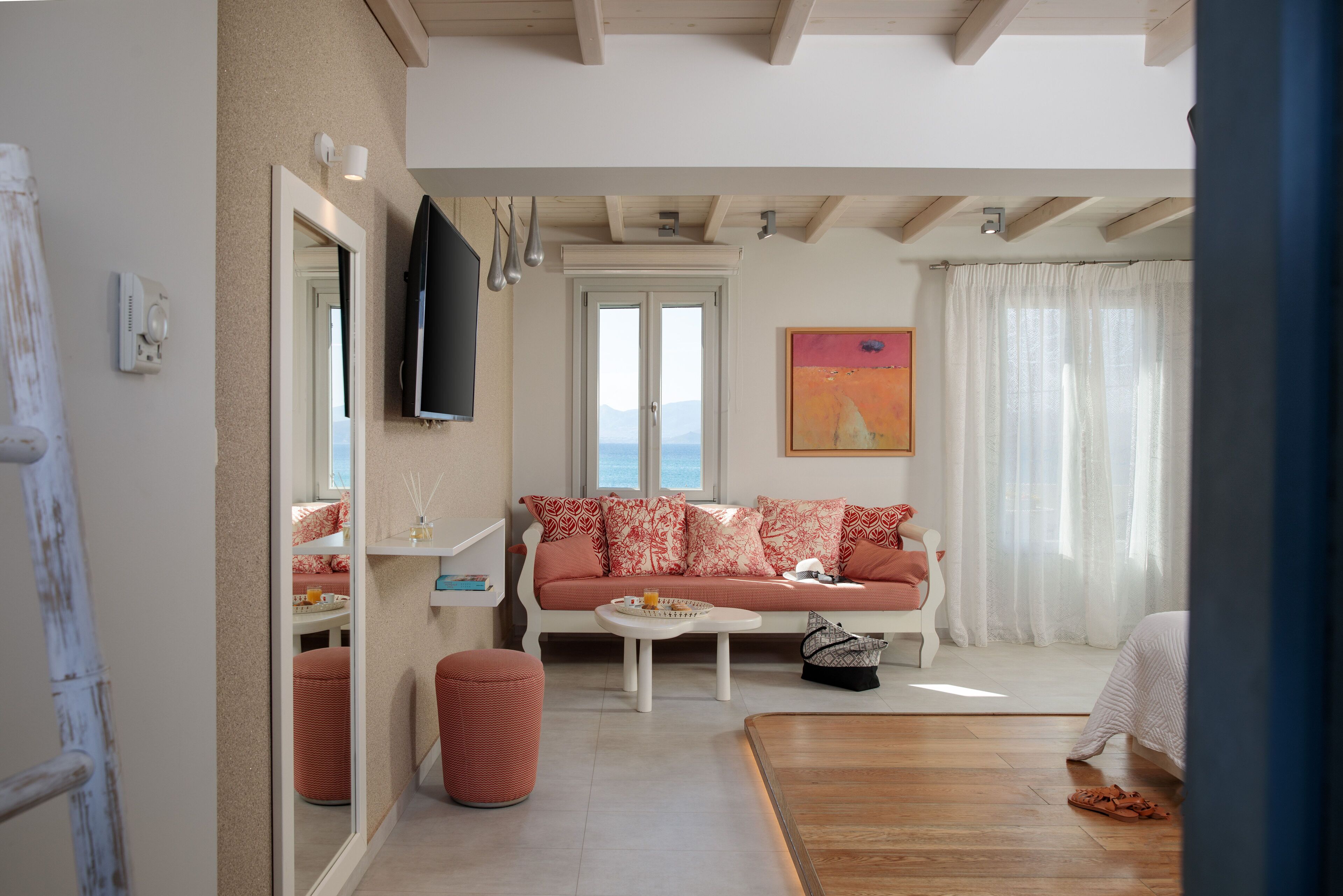 Ein Wohnzimmer mit Sofa, Hocker und Couchtisch. Ein Fernseher ist an der Wand montiert, ein Fenster mit Vorhängen und ein Gemälde hängt dort.