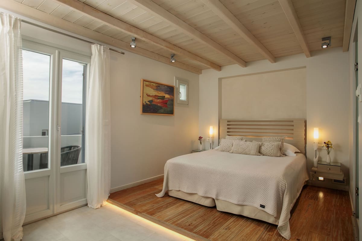 Een slaapkamer met een groot bed, houten vloer, een raam met gordijnen, een schilderij aan de muur en een nachtkastje met een lamp.