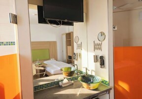 Habitación de hotel con cama, televisión, baño con dos lavamanos y espejo, y una mesita con caja de pañuelos.