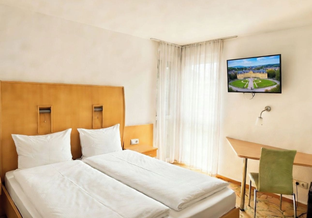 A hotel room with a double bed, a desk, a chair, a television, and a window with curtains.