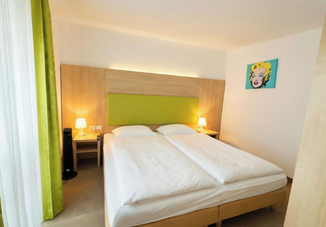 Ein Hotelzimmer mit Bett, Nachttischlampen, einem grünen Vorhang und einem Wandbild.