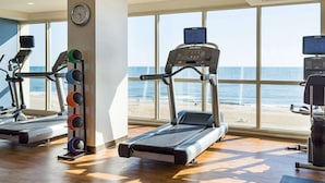 Une salle de sport équipée de tapis de course et d’appareils elliptiques, de grandes fenêtres donnant sur la plage et une horloge au mur.