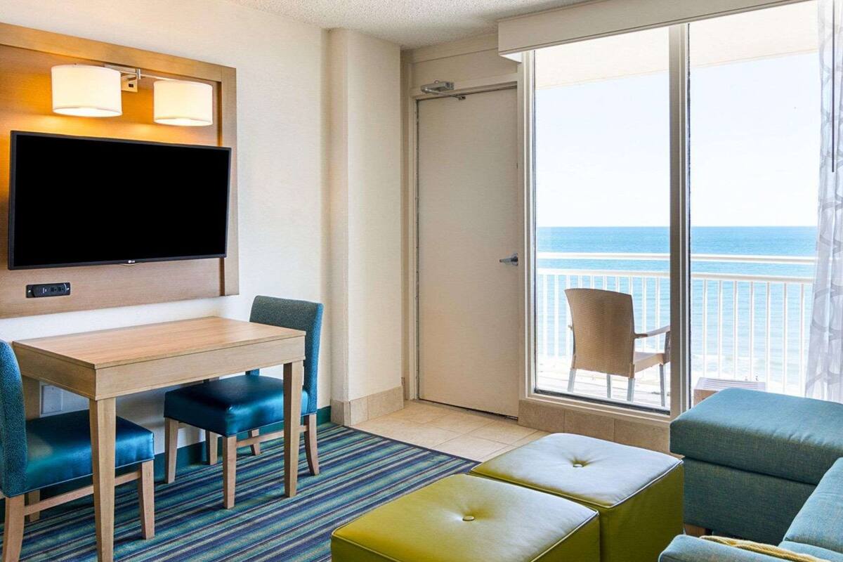Een hotelkamer met een televisie, een eettafel met stoelen, een balkon met een stoel en een bank.