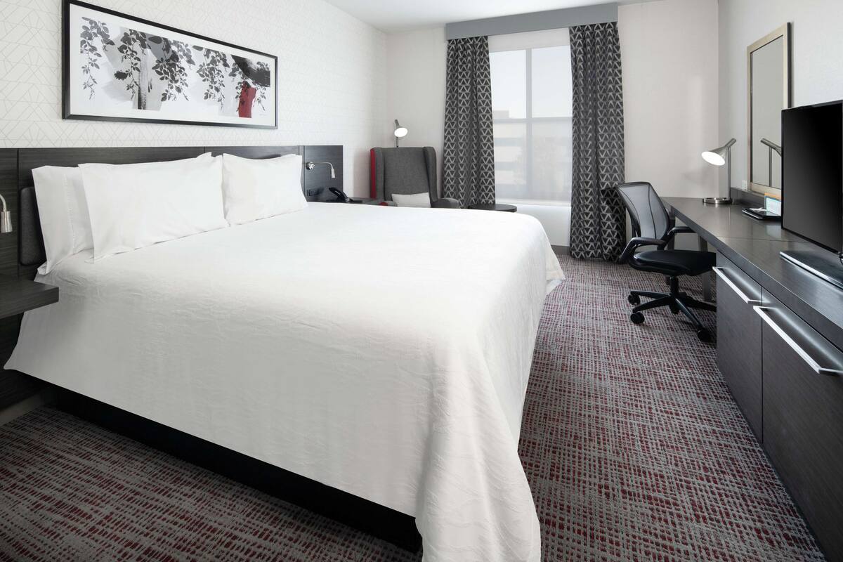 A hotel room with a large bed, a desk with a computer, a chair, a TV, and a phone on the nightstand.