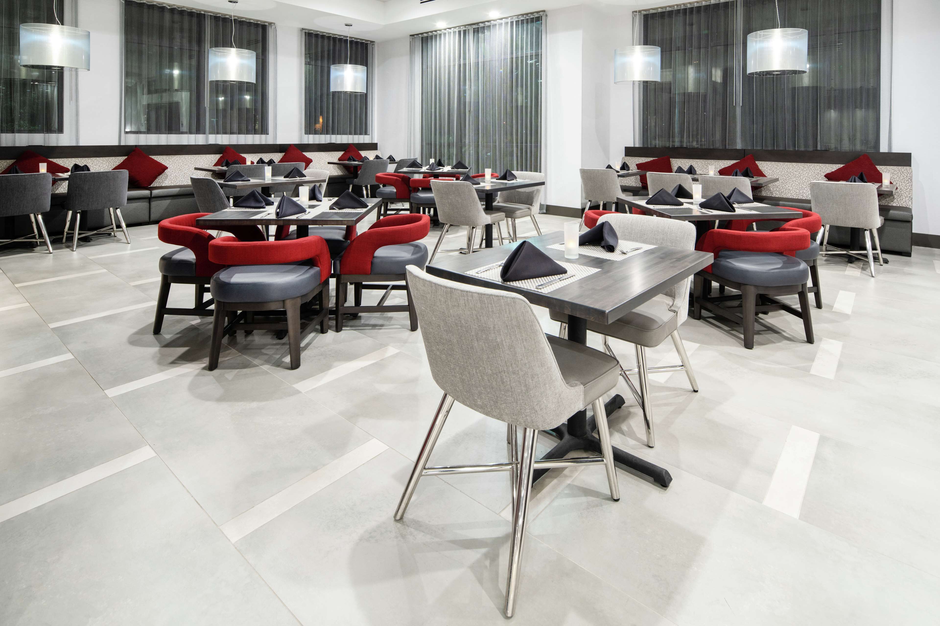 Un comedor moderno con varias mesas y sillas, ventanas amplias con cortinas, y un piso limpio cubierto de baldosas.