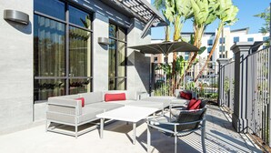An outdoor seating area with a sofa, chairs, and a table, a large umbrella, and a modern building with large windows and a metal fence. - Hilton Garden Inn Irvine Spectrum Lake Forest (Lake Forest)