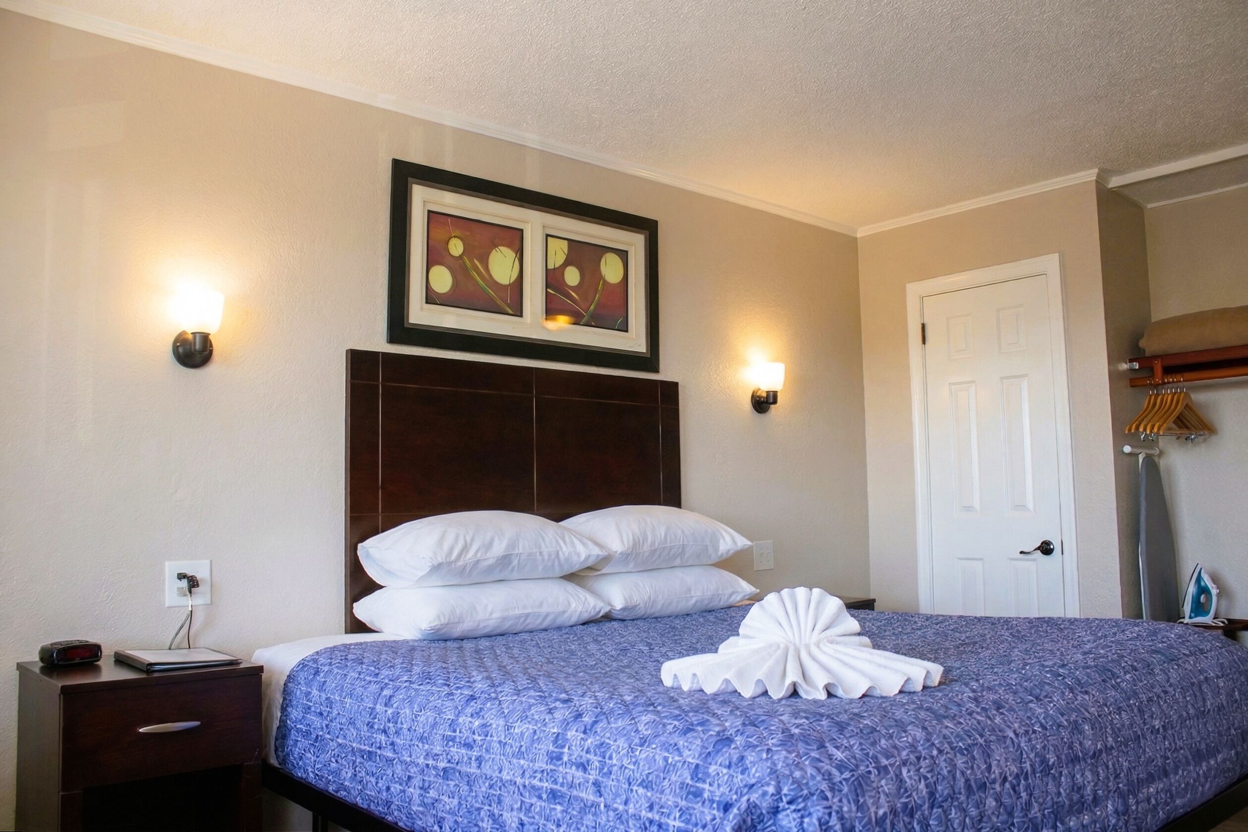 A hotel room with a bed, two wall-mounted lamps, a framed artwork, a white door, and a nightstand with a lamp and a clock.