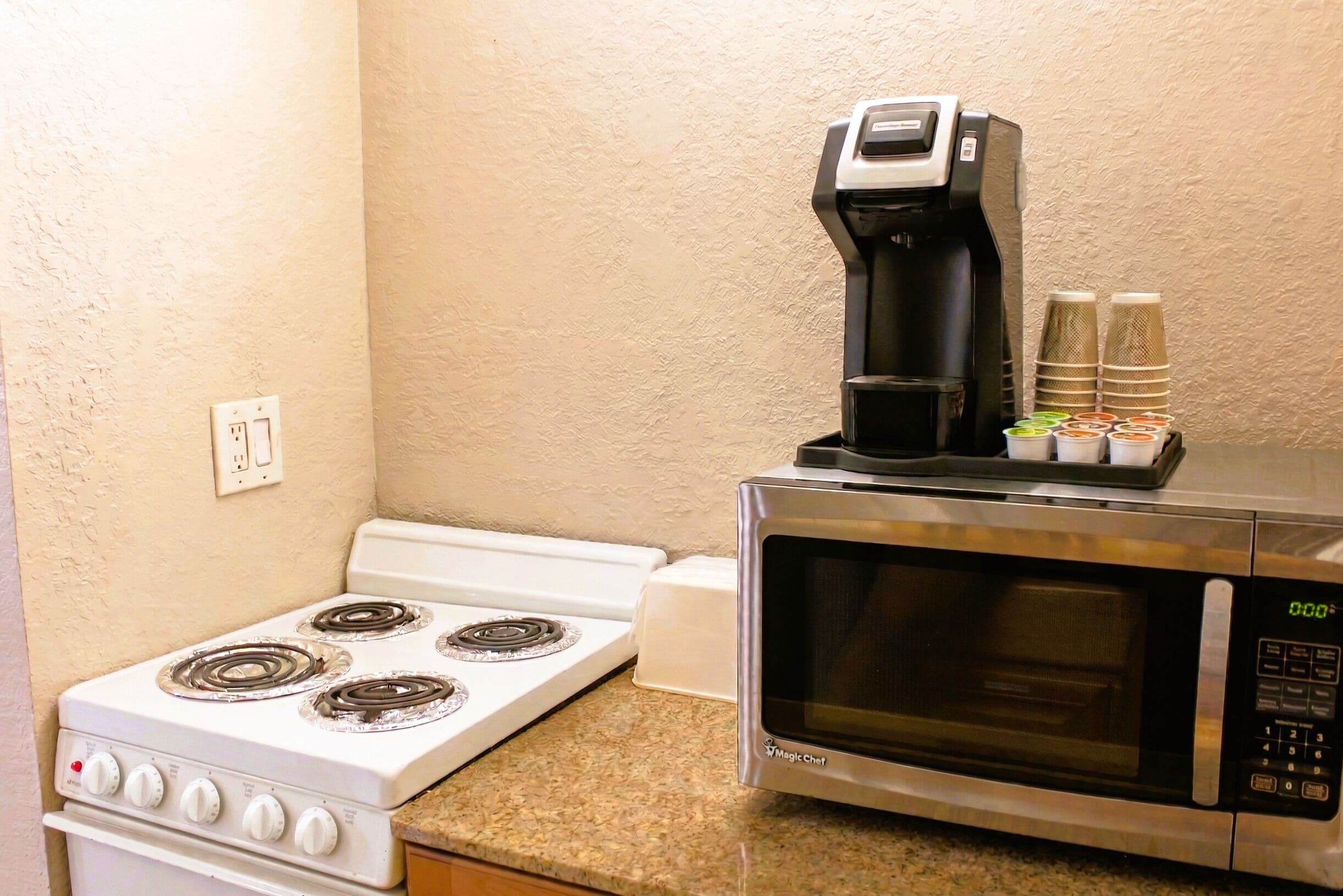 A kitchen with a stove, microwave, coffee maker, and stacked paper cups.