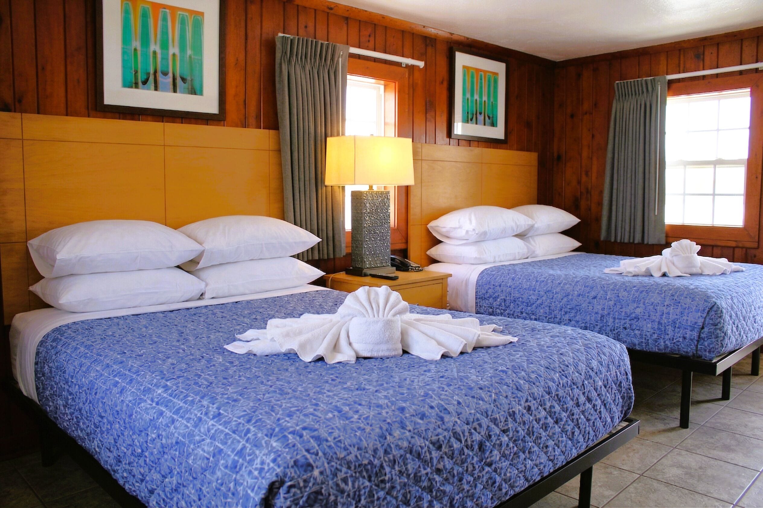 A hotel room with two beds, a nightstand with a lamp, wooden walls, and artwork on the walls.