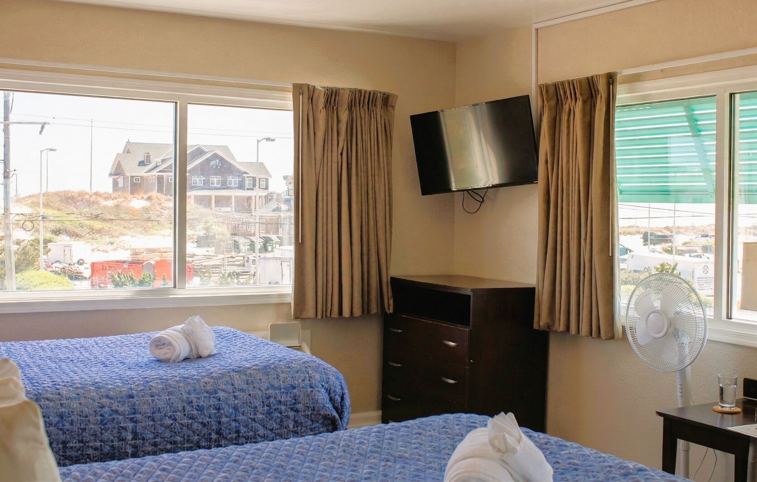 A hotel room with two beds, a TV, a dresser, a fan, and a window with a view of buildings and a street.