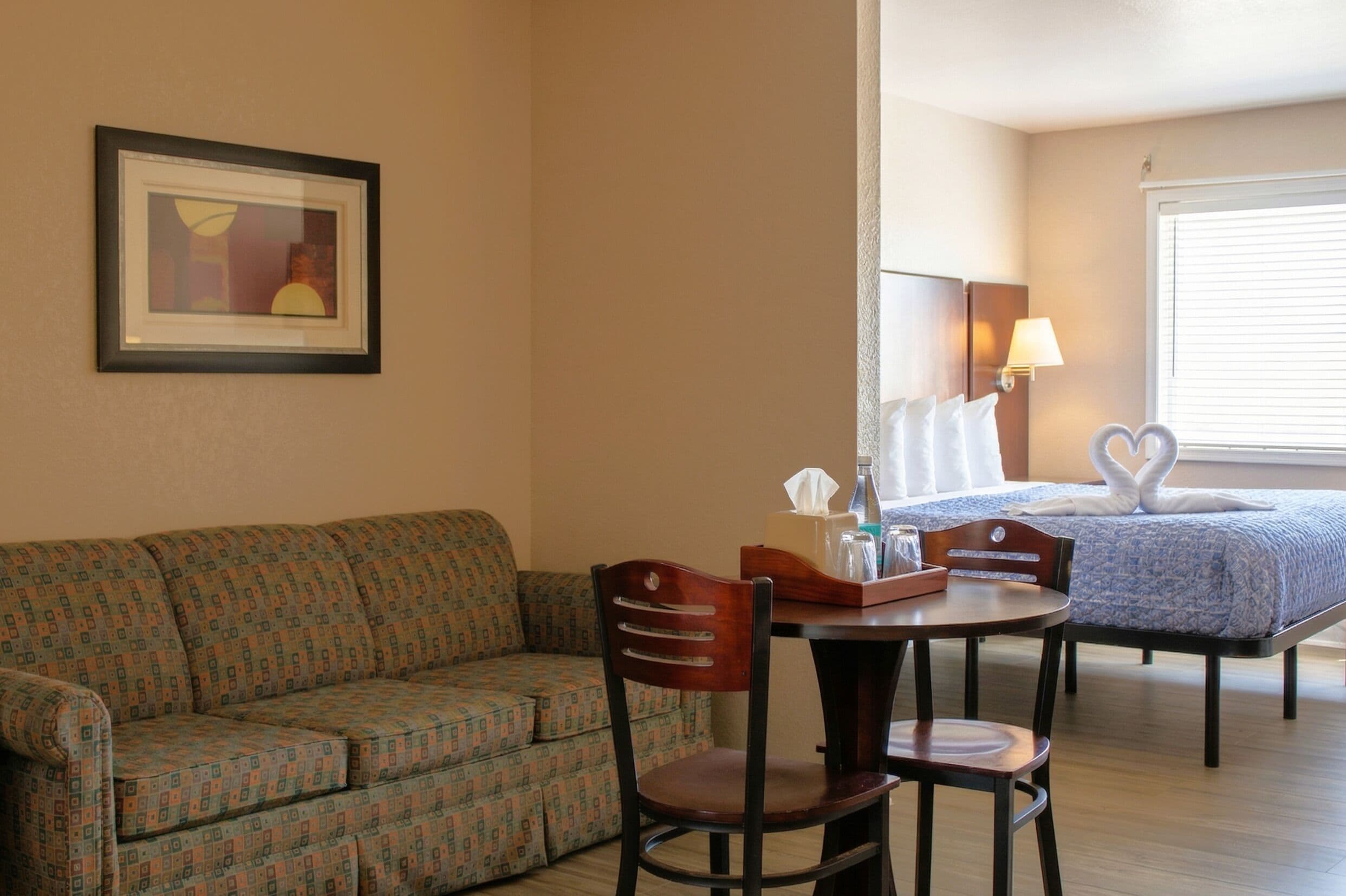 A room with a patterned sofa, a dining table with chairs, a framed picture on the wall, and a bed with heart-shaped pillows.