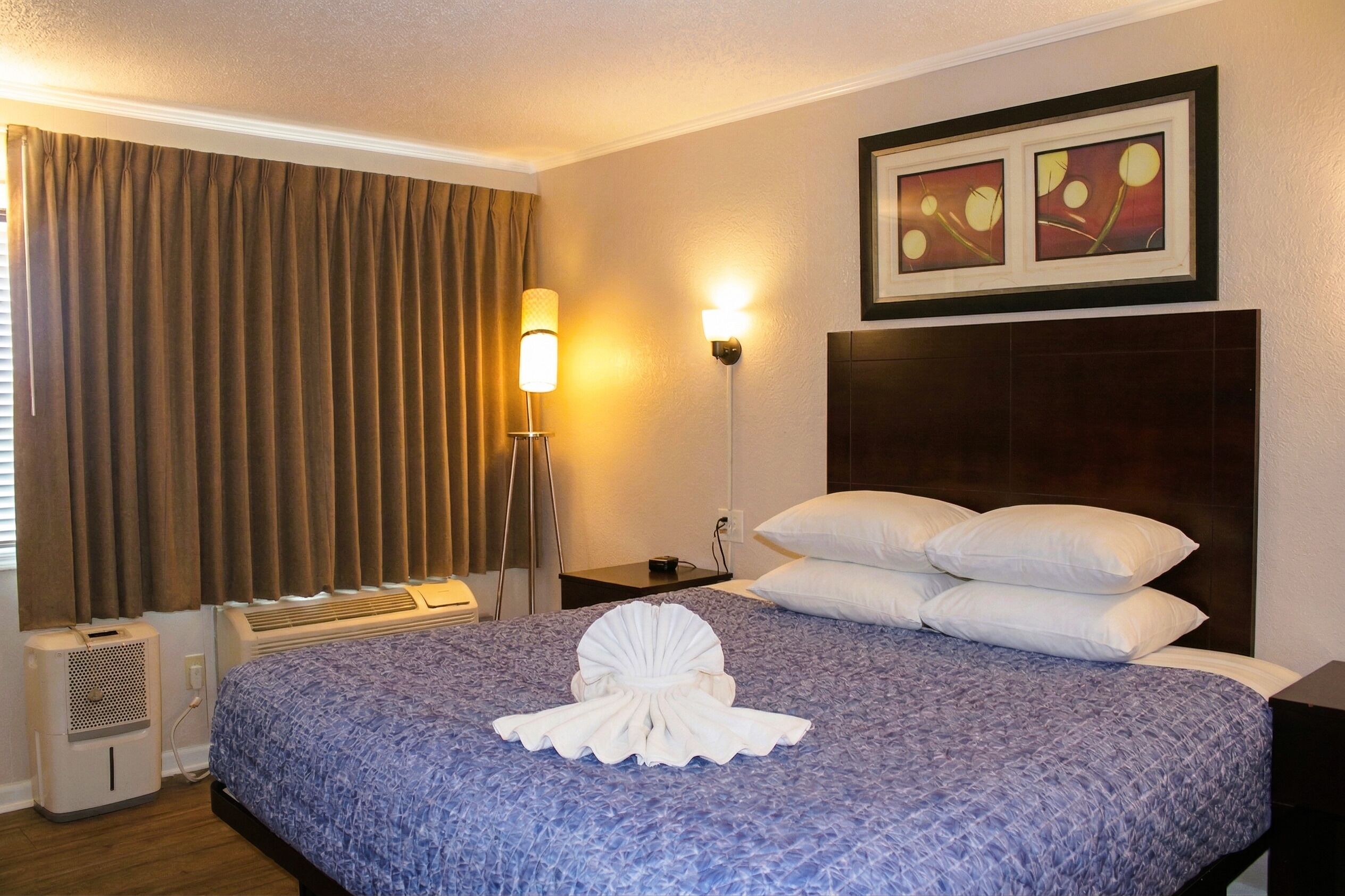 A hotel room with a bed, a headboard, a wall-mounted picture, a bedside lamp, and a window with curtains.