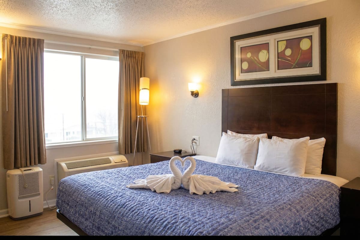 Ein Hotelzimmer mit einem großen Bett, einem Wandbild, einem Nachttisch und einem Fenster mit Vorhängen.