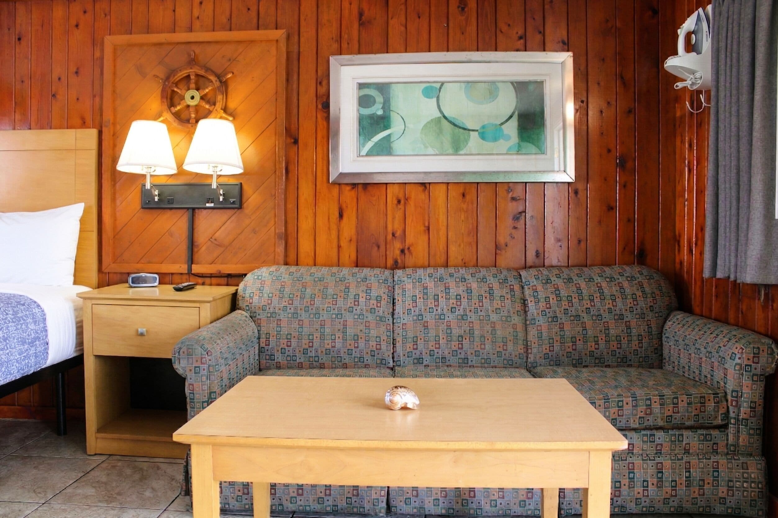 Une pièce aux murs lambrissés, comprenant un lit, une table de chevet avec une lampe, un canapé à motifs et un tableau abstrait encadré.