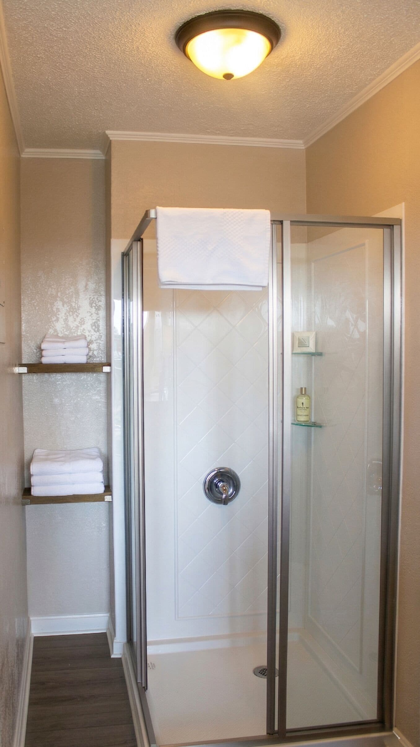 A glass-enclosed shower with a white towel on the door, a shelf with folded towels, and a bottle of shampoo on the wall.