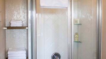 A glass-enclosed shower with a white towel on the door, a shelf with folded towels, and a bottle of shampoo on the wall.