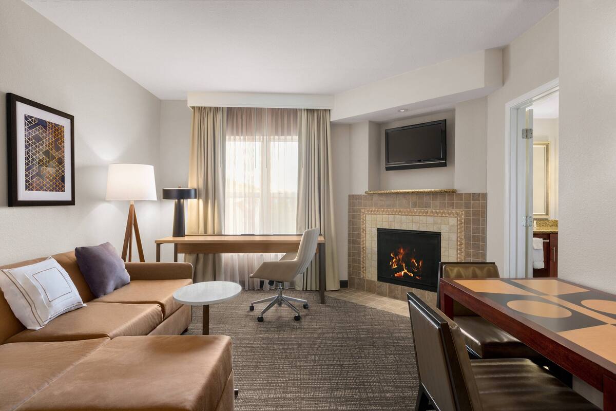 A hotel room with a sofa, a dining table, a fireplace, a TV, and a desk with a lamp.
