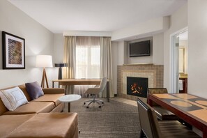 A hotel room with a sofa, a dining table, a fireplace, a TV, and a desk with a lamp.