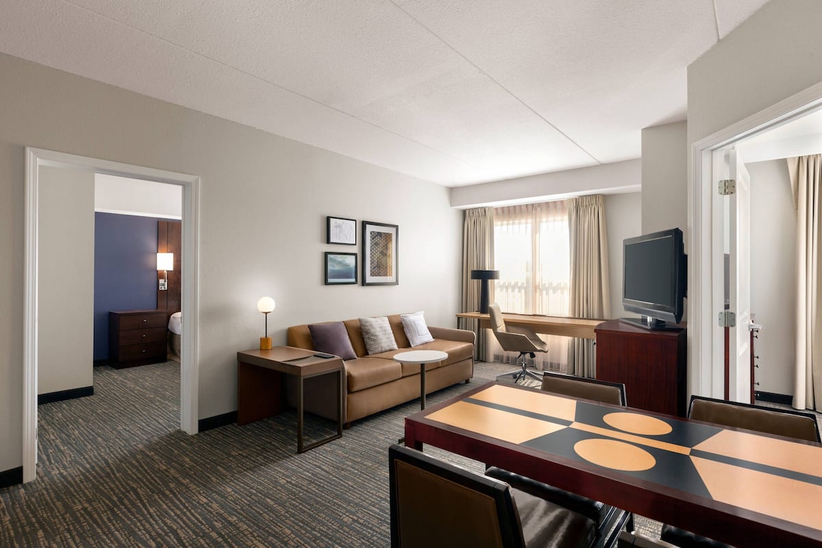 A hotel room with a sofa, a small table, a TV, and a dining area with chairs.