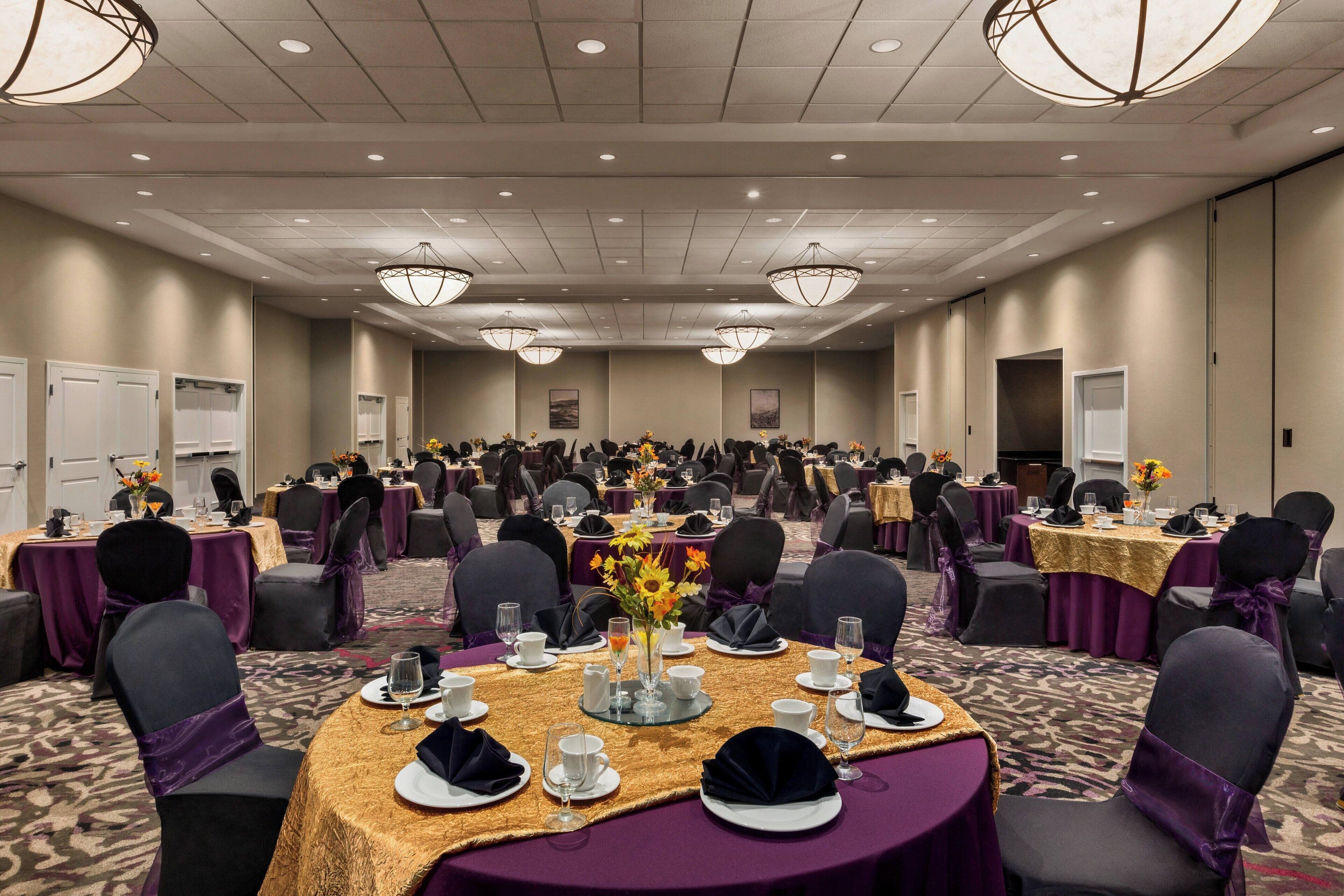 A banquet hall with round tables set for a formal event, featuring gold tablecloths and purple chair covers.