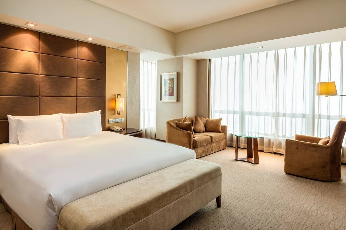 A hotel room with a large bed, a sofa, a small table, and a large window with curtains.