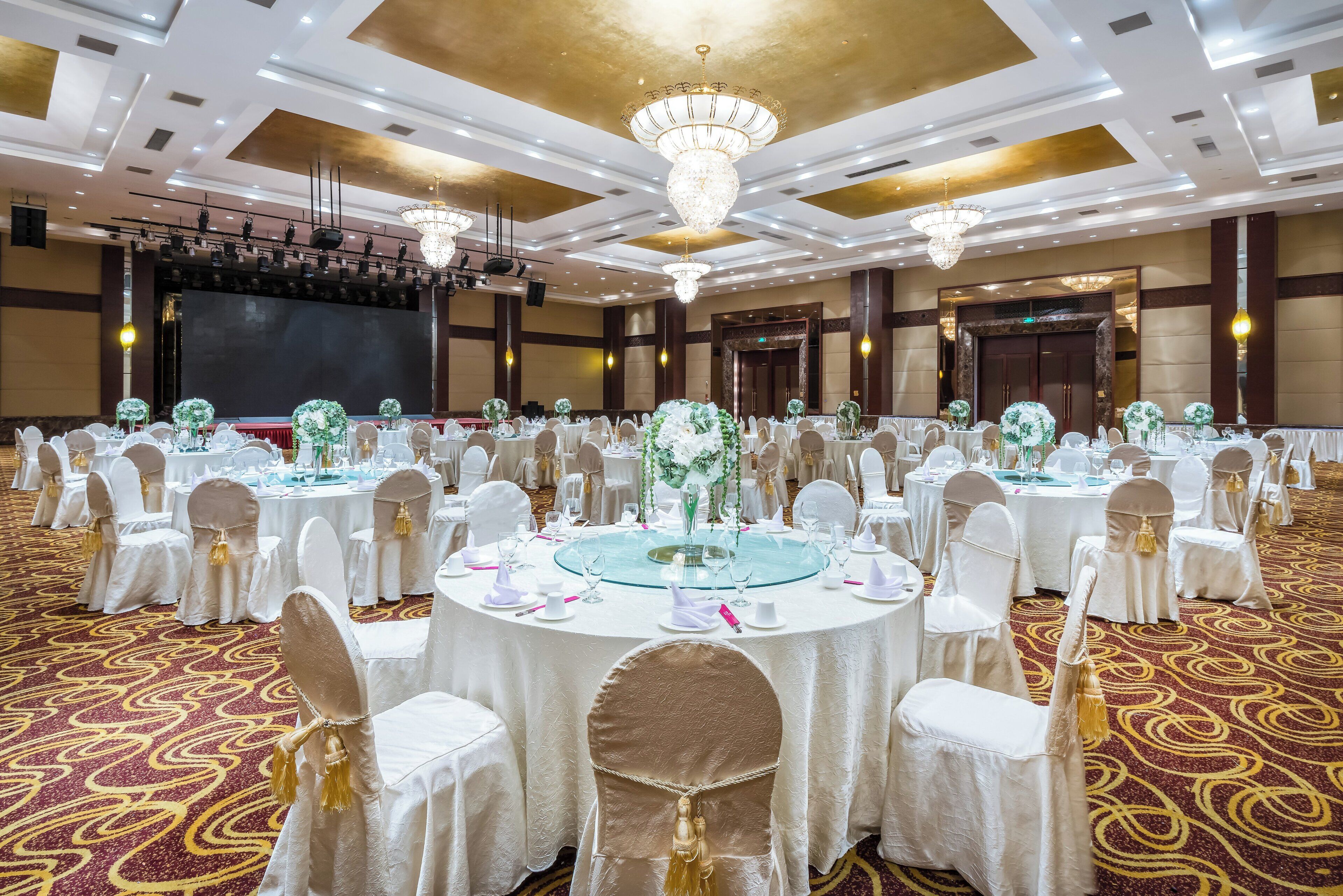 Un salón de banquetes con mesas redondas, manteles blancos y sillas con cintas doradas.