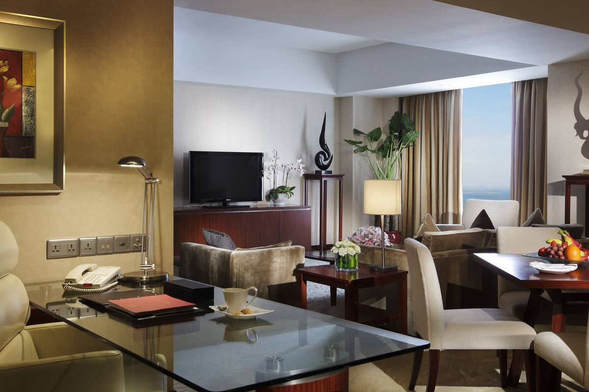 A hotel room with a glass coffee table, a sofa, a TV, a desk with a lamp, a plant, and a view of the city.