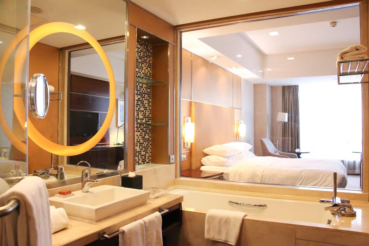 A modern hotel bathroom with a large mirror, two sinks, a bathtub, and a visible bed in the adjacent room.