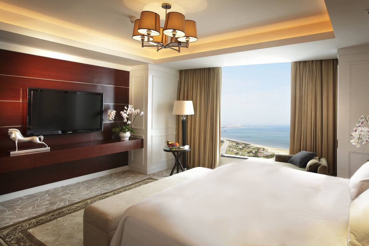 A hotel room with a large bed, a flat-screen TV mounted on the wall, a small table with a lamp, and a view of the sea.