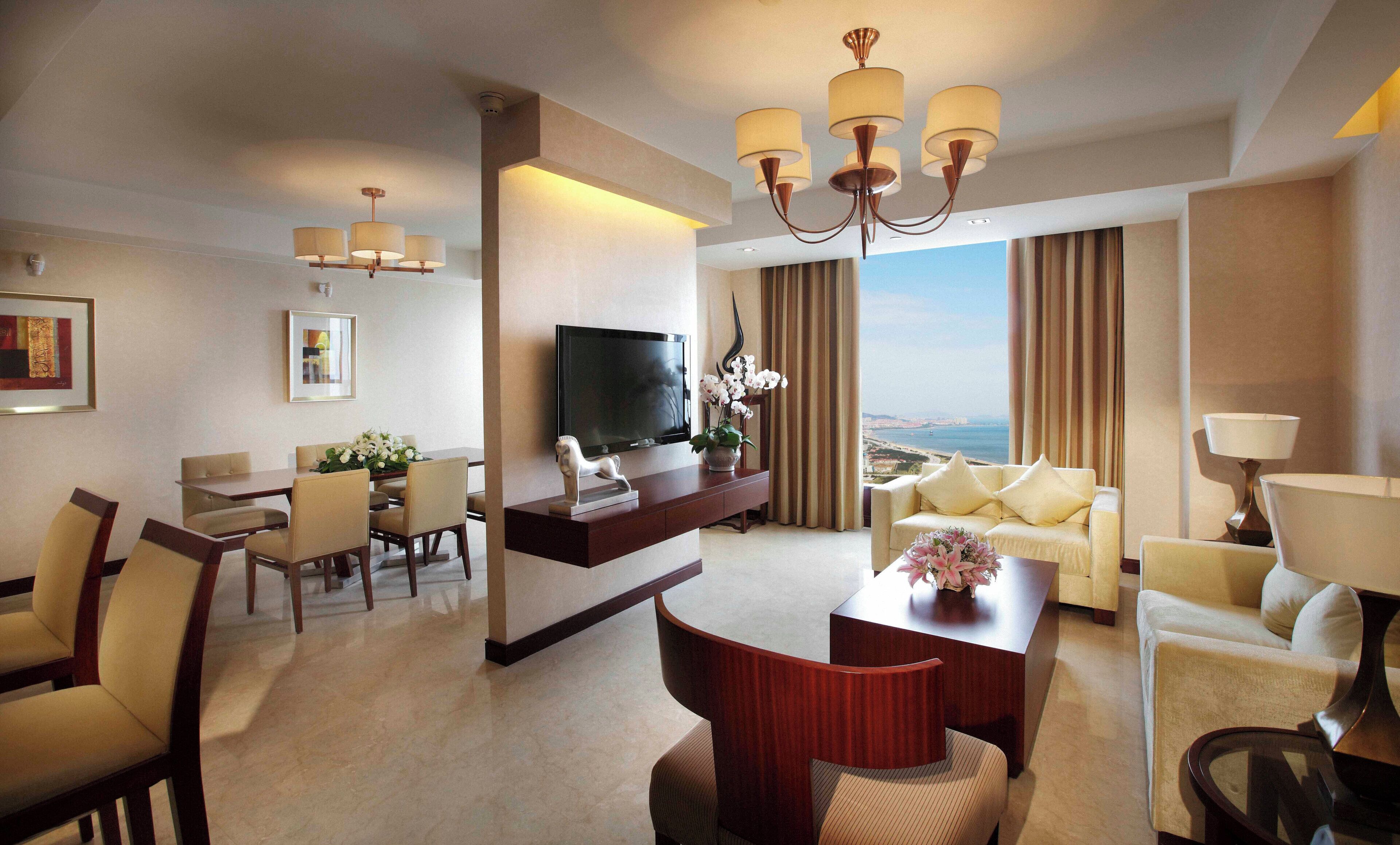 A hotel room with a TV, seating area, dining table, and a view of the sea.