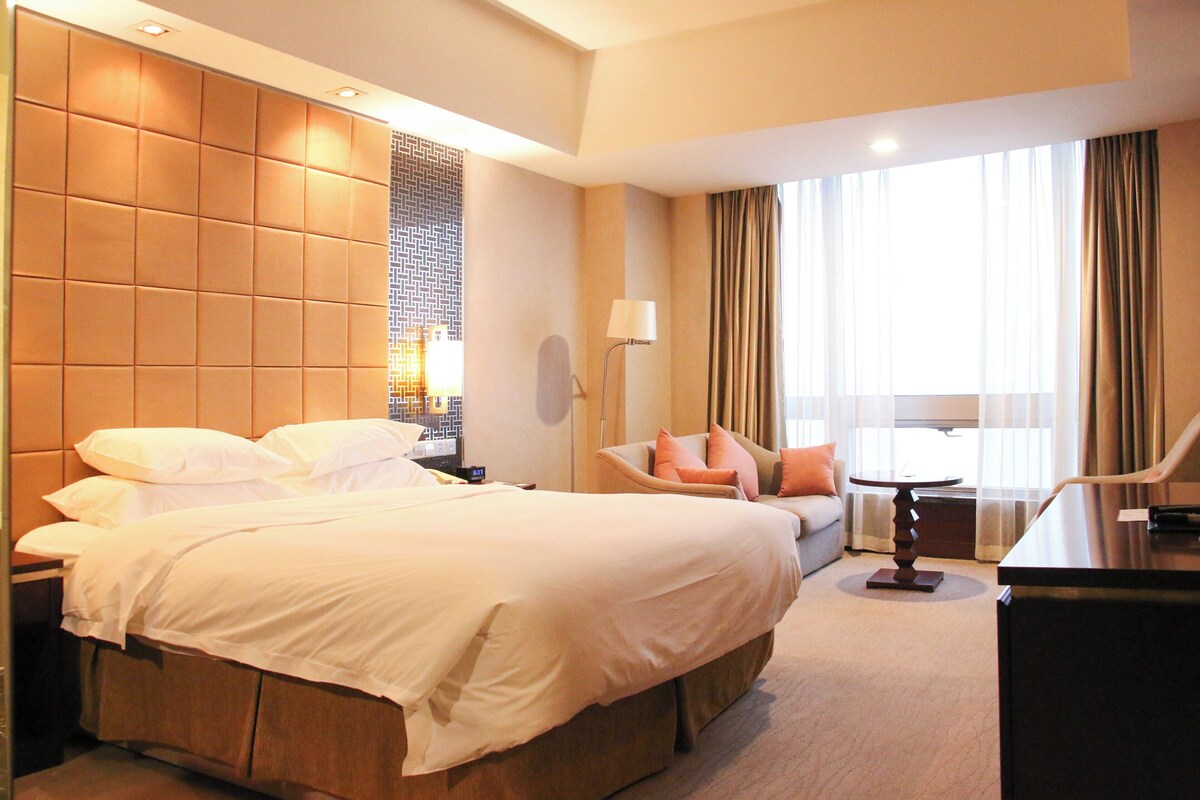 A hotel room with a large bed, a desk, a chair, a lamp, and a window with curtains.