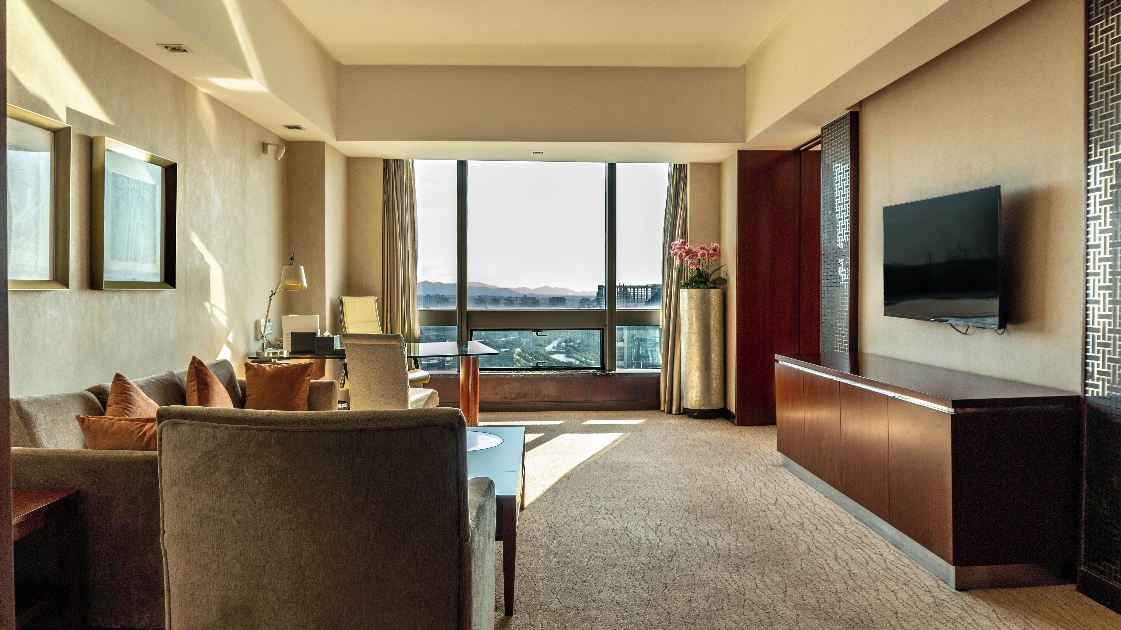 A hotel room with a large window, a flat-screen TV, a desk with a laptop, and a seating area with a sofa and armchairs.