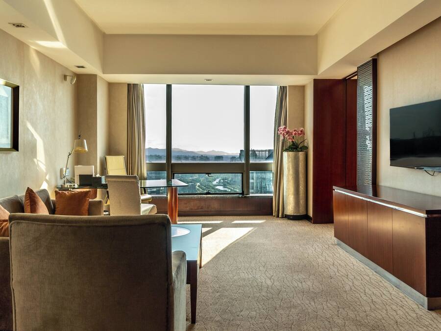 A hotel room with a large window, a flat-screen TV, a desk with a laptop, and a seating area with a sofa and armchairs.