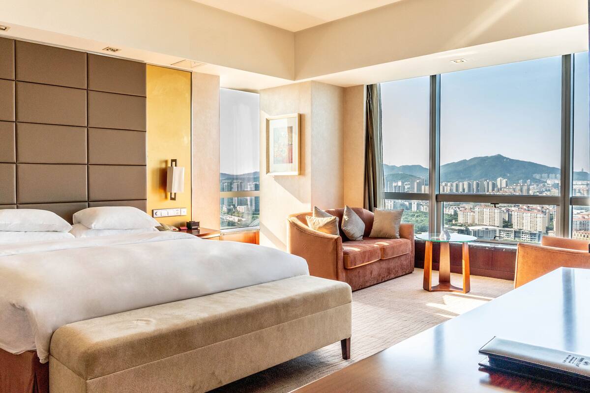 A hotel room with a large bed, a sofa, a desk, and a city view through large windows.