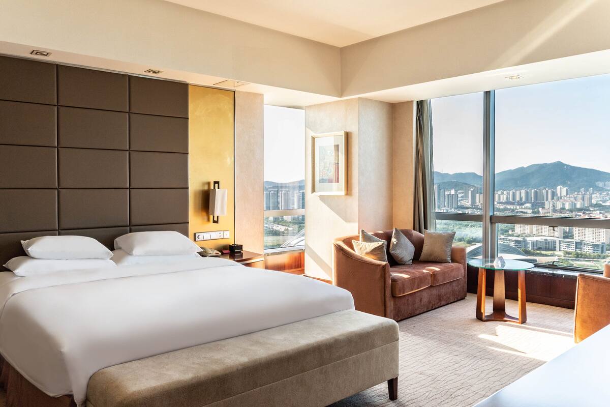 A modern hotel room with a large bed, a seating area with a view of the city, and a balcony with a cityscape view.
