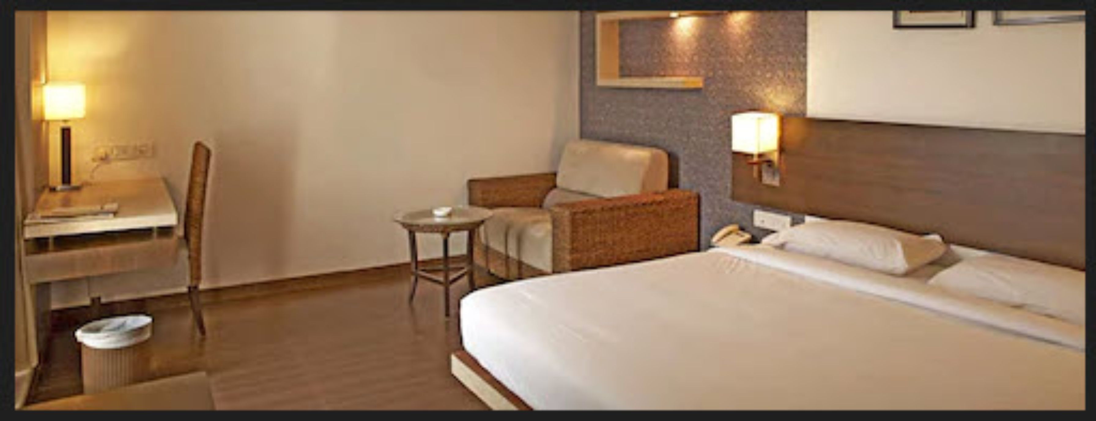 A hotel room with a bed, a desk, a chair, a small table, and two wall-mounted lamps.
