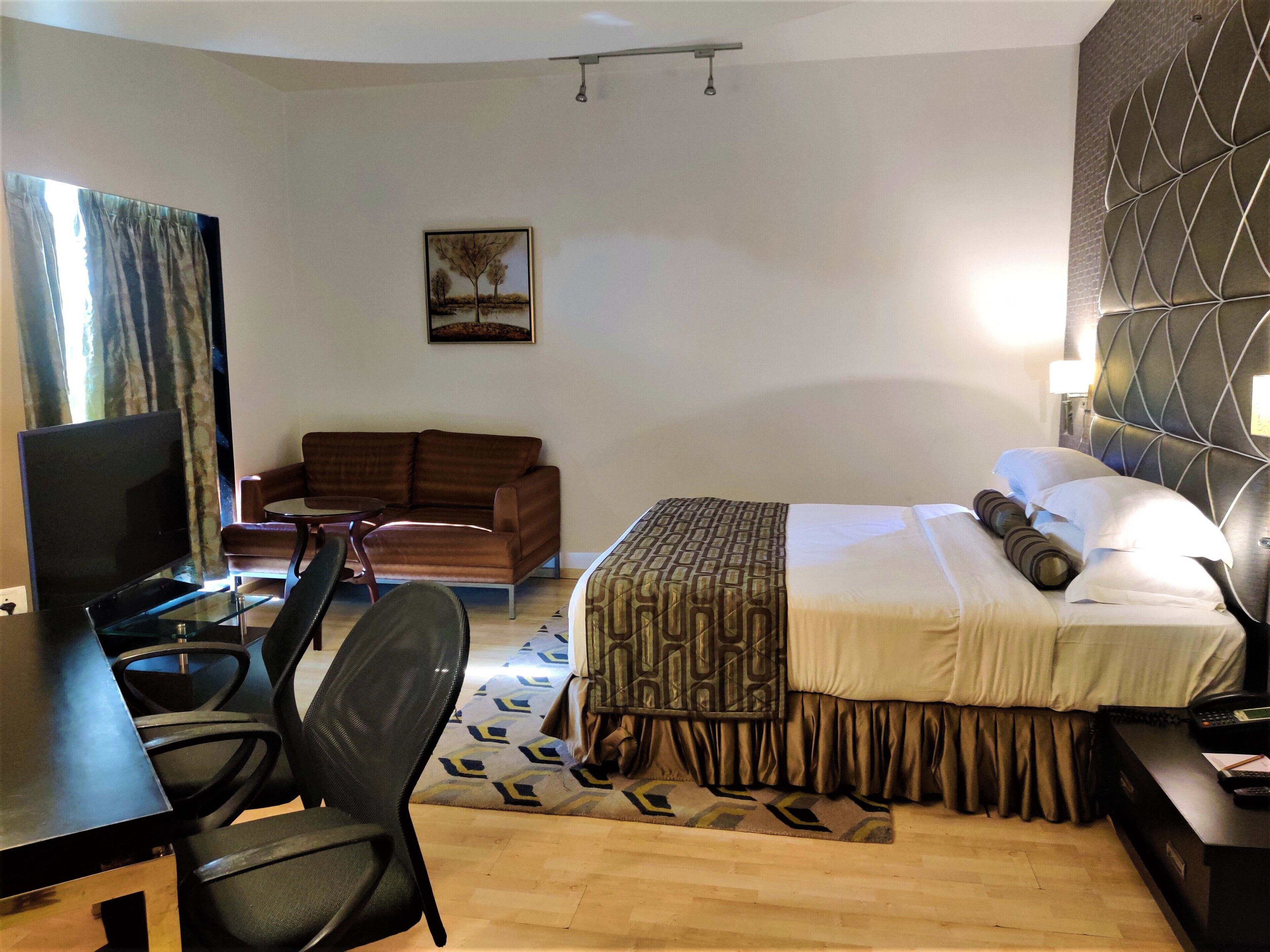 A hotel room with a bed, a desk with a computer, a TV, a sofa, and a painting on the wall.