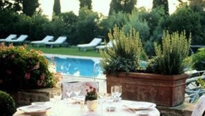 An outdoor dining area with a round table set for two, surrounded by greenery and a pool in the background. - Hotel Satkar Residency (Thane)