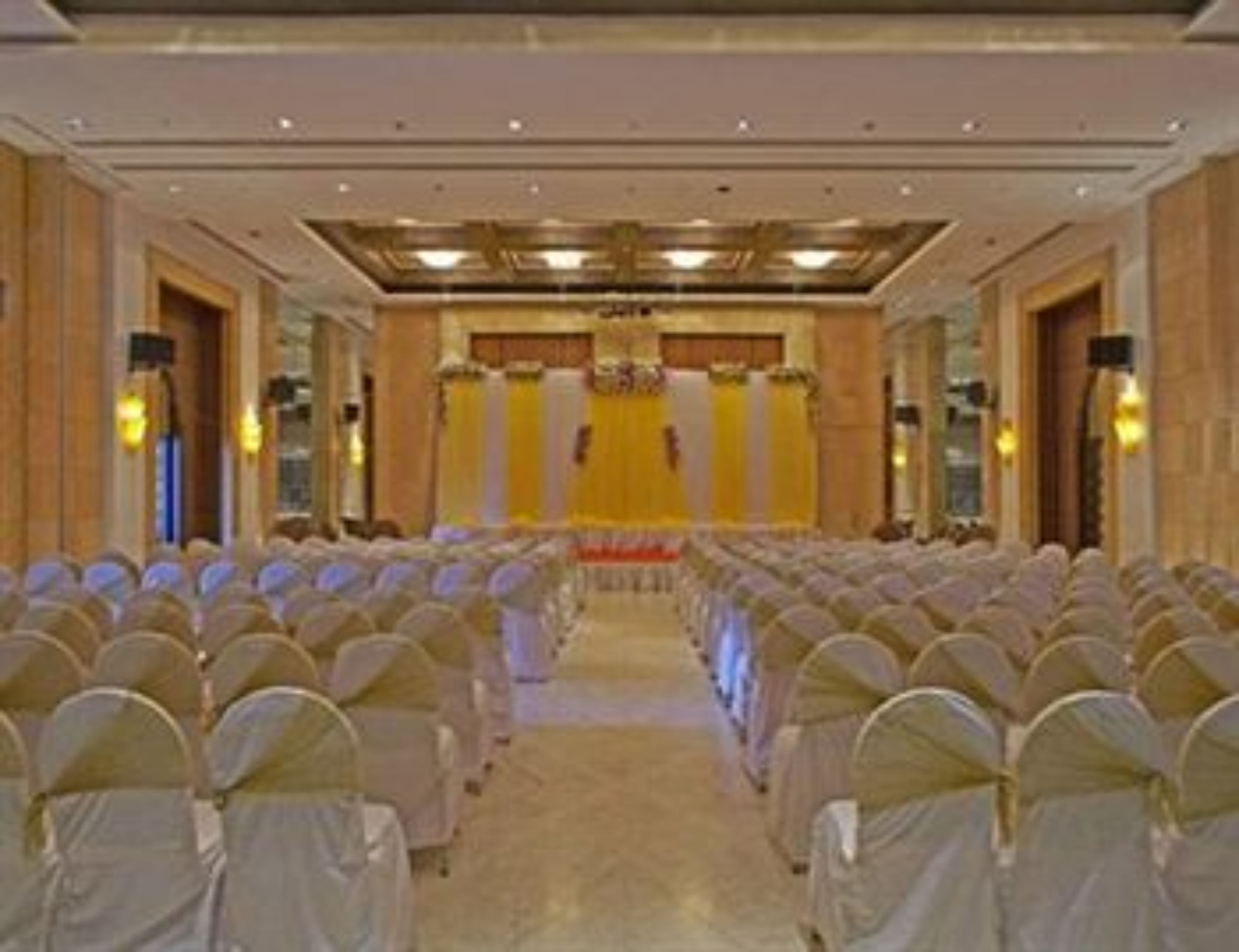 A hotel conference room with rows of white chairs, a stage with a yellow backdrop, and chandeliers.