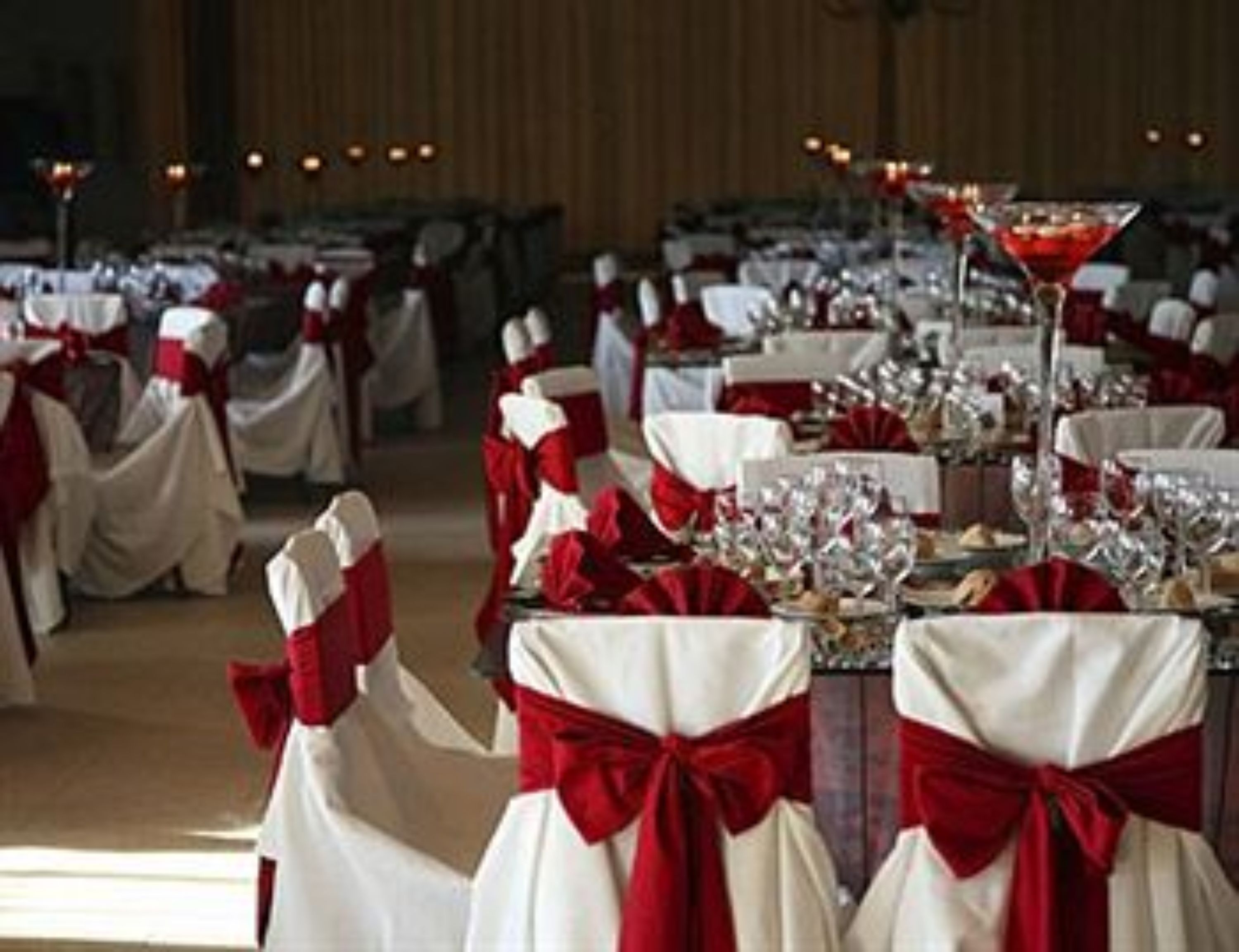 Un salón de banquetes con mesas dispuestas para un evento formal, cada una con un camino de mesa rojo y blanco y una copa de vino tinto.