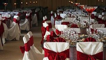 Un salón de banquetes con mesas dispuestas para un evento formal, cada una con un camino de mesa rojo y blanco y una copa de vino tinto.